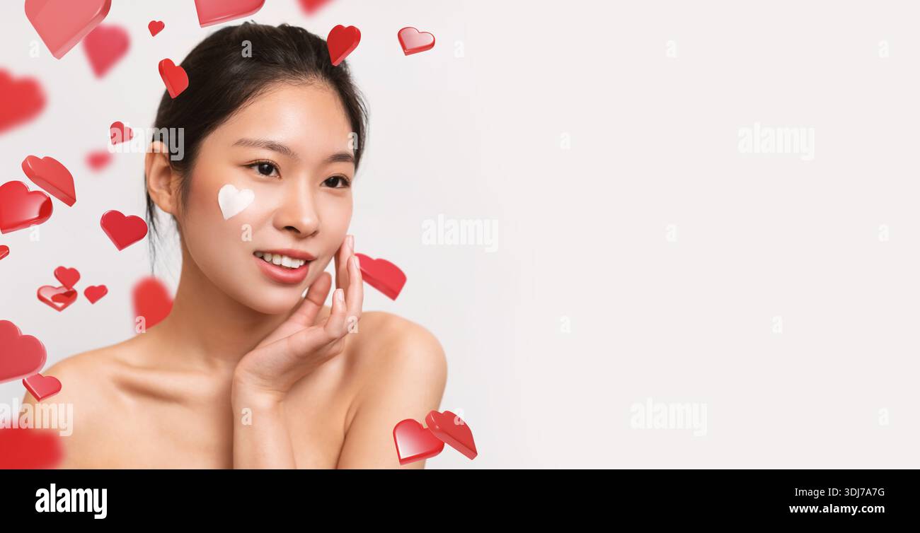 Asiatische Frau zeigt herzförmige Creme auf der Wange, während sie drinnen von weißem Hintergrund posiert Stockfoto