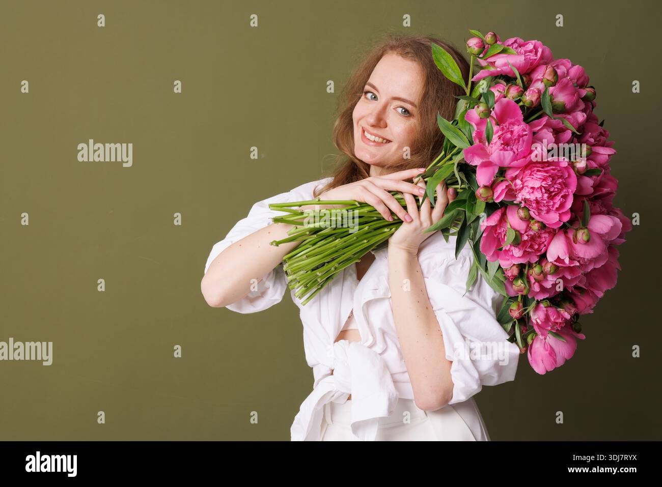 Großer Strauß rosafarbener Pfingstrosen, gehalten von einer Frau in weiß vor olivgrünem Hintergrund. Stockfoto