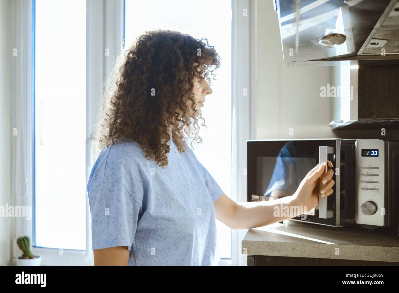 Eine Frau steht in einer Küche an einer Mikrowelle. Sie bereitet sich darauf vor, Essen zu erhitzen. Sonnenlicht tritt durch das Fenster ein. Die Küche ist ordentlich und einfach eingerichtet. Stockfoto