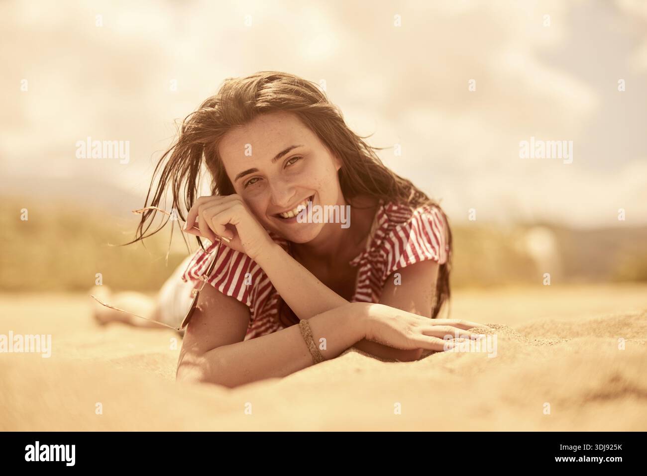 Nahaufnahme einer lächelnden Frau, die entspannt auf dem Sand liegt und die sonnige Strandatmosphäre genießt, unbeschwerte Momente, Sonnenschein, exhuberantes Lächeln genießt, Stockfoto