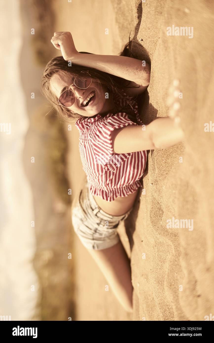 Frau liegend auf dem Sand, lächelnd, in lässigem gestreiftem Hemd und Sonnenbrille am Strand, fröhliches Glück, überglücklich sein, Entspannung, Wellness, holi Stockfoto