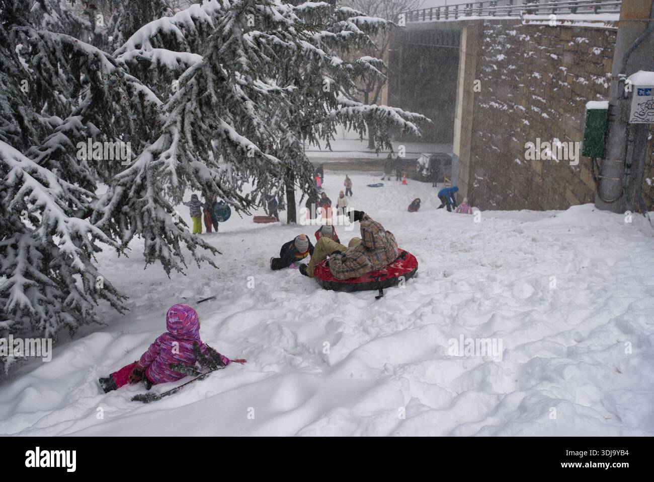 Eine schneebedeckte Piste mit Kinderrodeln und Tubing. Schnee fällt, während Menschen unterschiedlichen Alters Wintererholung in der Nähe einer Steinmauer und Brücke genießen. Stockfoto