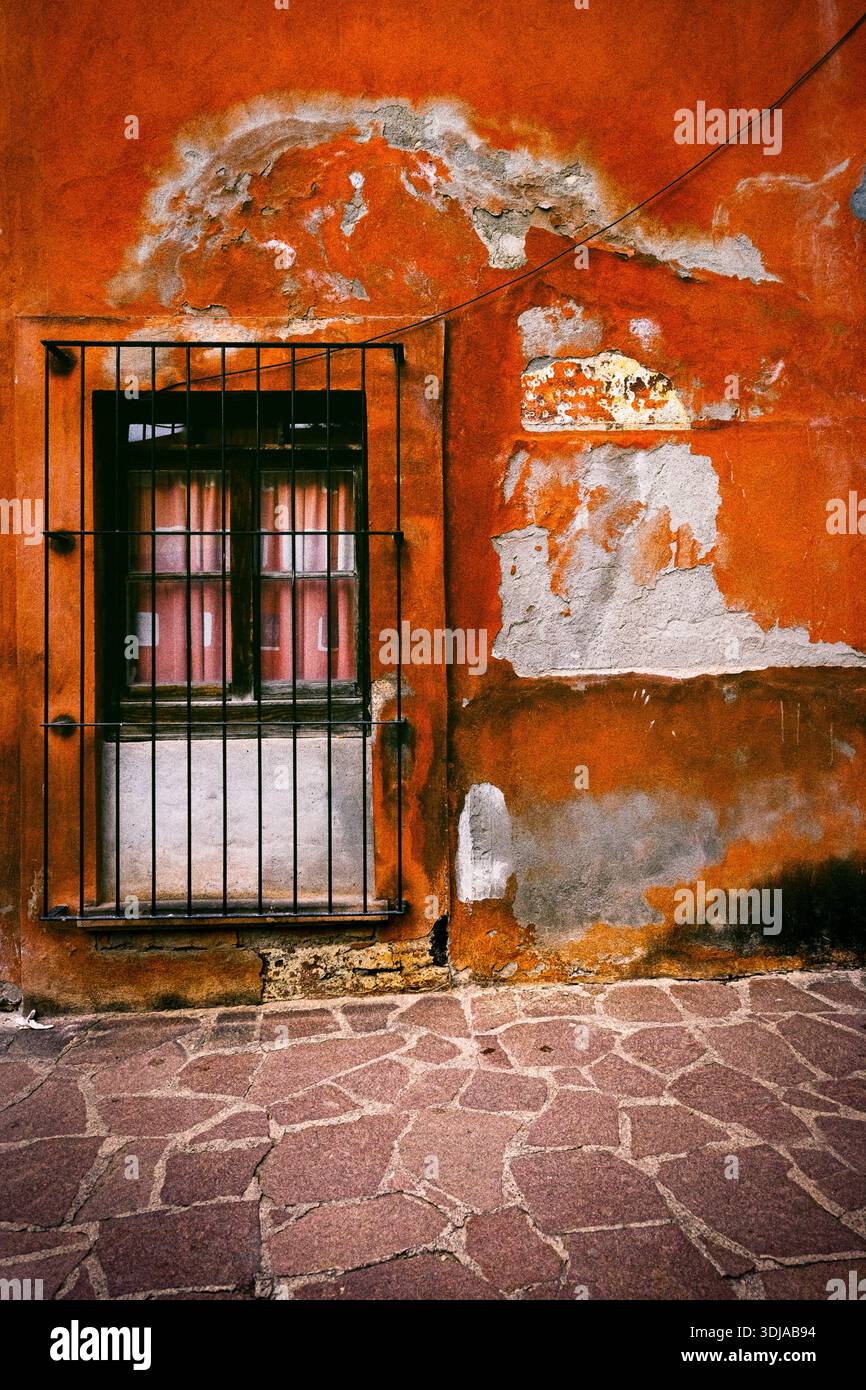 Eine orangefarbene Wand mit abblätternder Farbe zeigt ein vergittertes Fenster, das durch transparente Vorhänge Anklänge eines Innenraums offenbart Stockfoto