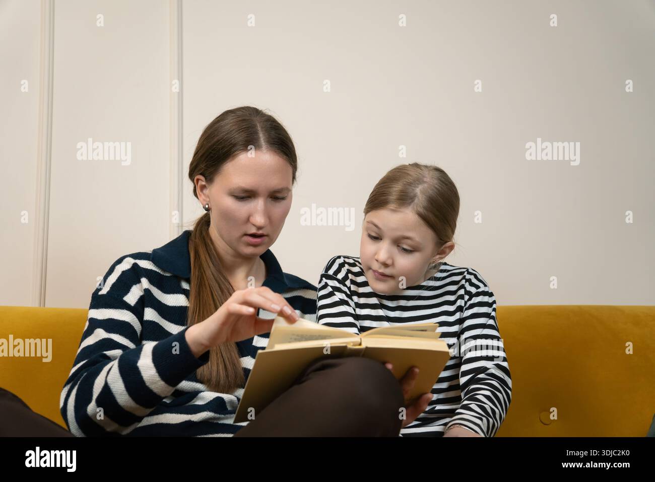 Mutter und Tochter verbinden sich beim Lesen eines Buches, sitzen zusammen auf einem gelben Sofa zu Hause, nehmen an einem friedlichen gemeinsamen Lernerlebnis Teil und enjo Stockfoto