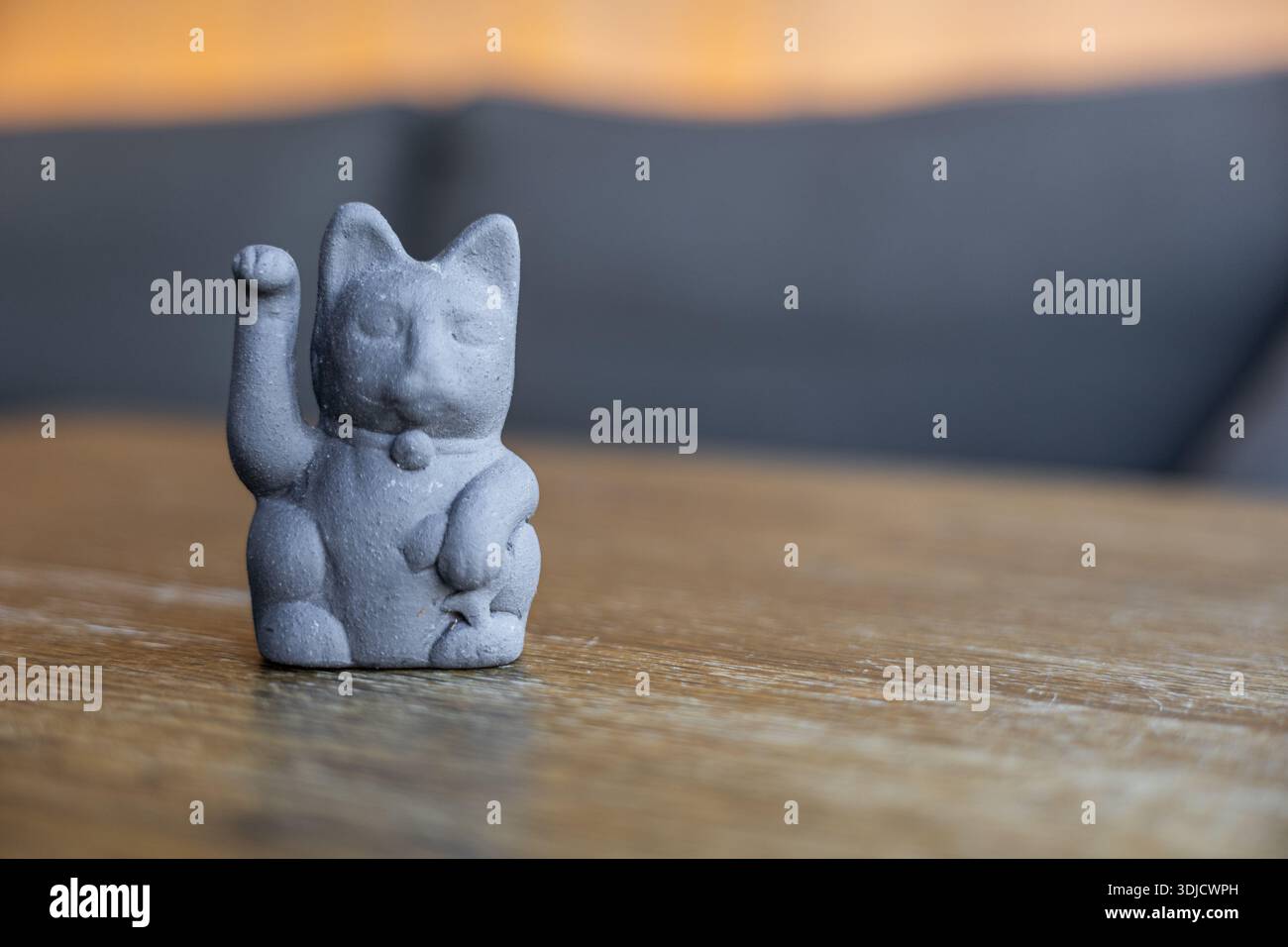 Eine kleine graue Keramikkatzenfigur mit einer erhobenen Pfote, die auf einem Holztisch sitzt. Der Hintergrund ist sanft verschwommen und hat einen warmen, neutralen Ton. Stockfoto
