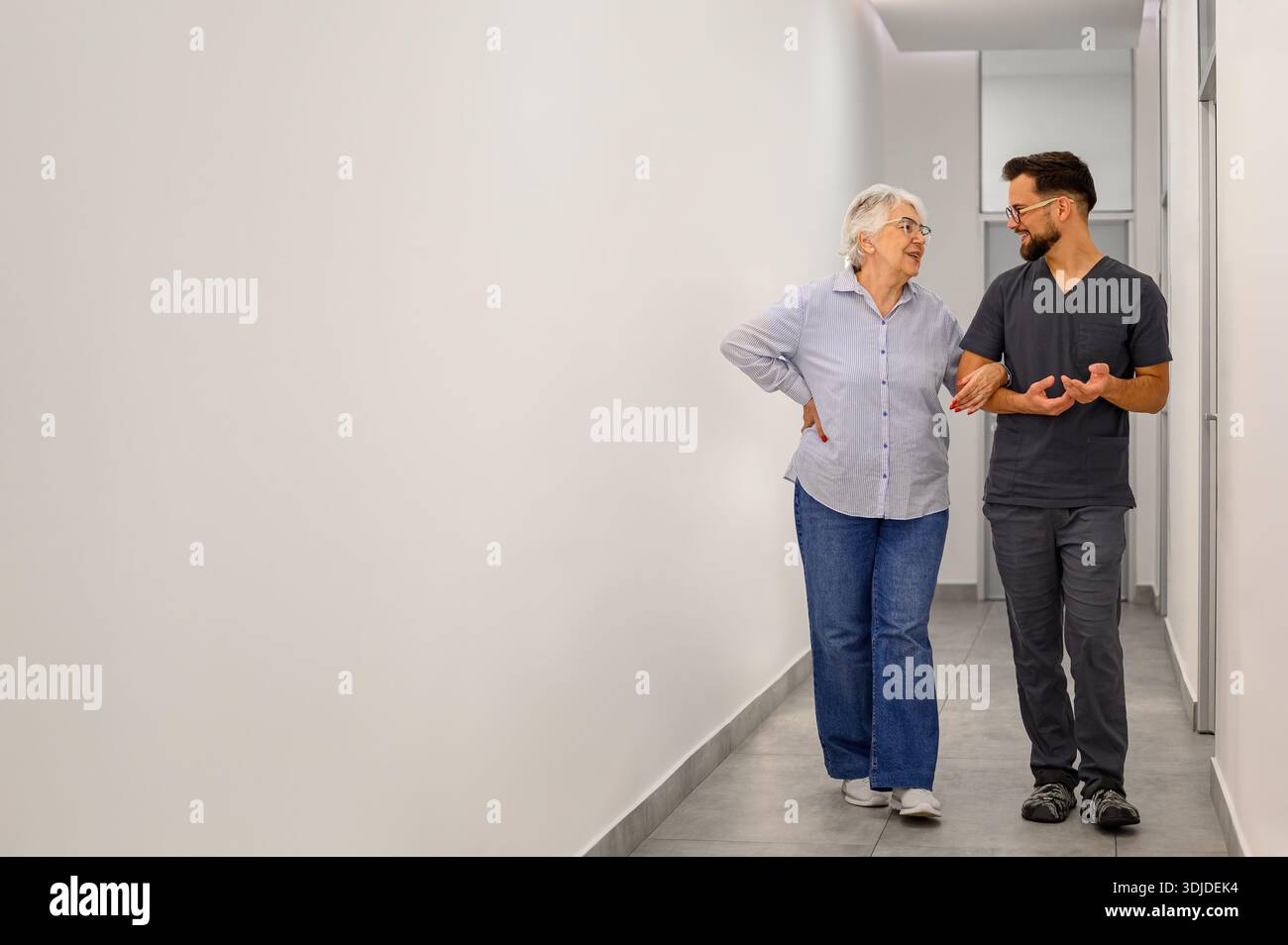 Volle Länge der älteren Frau, die mit dem männlichen Arzt Arm in Arm geht, während sie Symptome und Behandlung in der Krankenhaus-Lobby bespricht Stockfoto