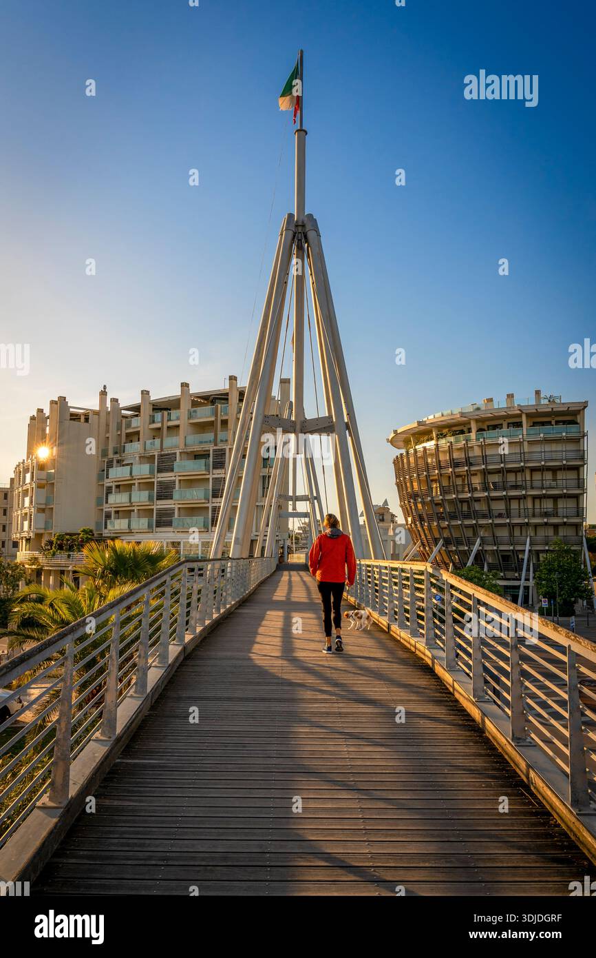 Einsame Frau geht mit ihrem Hund im warmen Licht des Sonnenaufgangs auf der Ponte della Valletta. Rimini, Emilia Romagna, Italien, Europa. Stockfoto