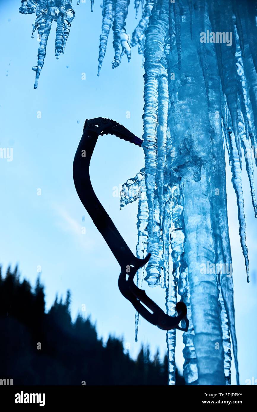 Nahaufnahme des Eisaxt-Werkzeugs zum Klettern mit scharfem Schnabel an der gefrorenen Eiswand großer Wasserfall gegen blauen Ski in den Winterbergen. Sportbergsteigen Stockfoto