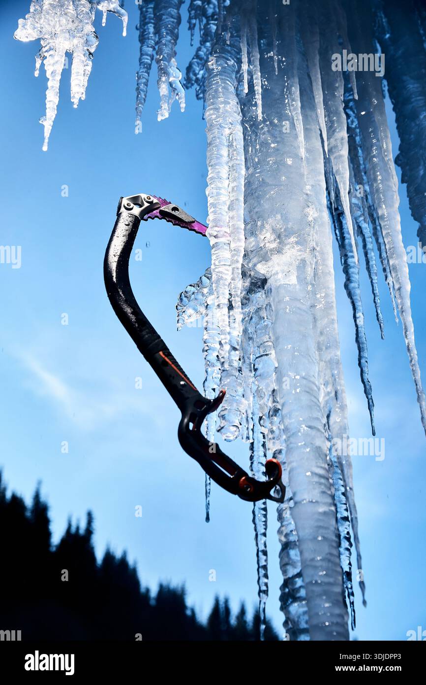 Nahaufnahme des Eisaxt-Werkzeugs zum Klettern mit scharfem Schnabel an der gefrorenen Eiswand großer Wasserfall gegen blauen Ski in den Winterbergen. Sportbergsteigen Stockfoto