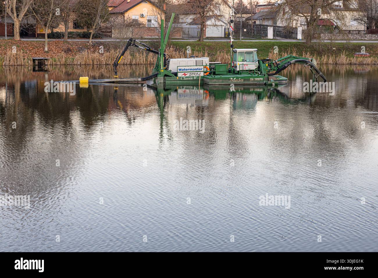 VINKOVCI, KROATIEN, 12.27.2025: Ein grünes Arbeitsboot mit hydraulischem Baggerarm fährt auf einem ruhigen See in der Nähe eines Wohngebietes. Maschinen- und Wasserreferenz Stockfoto