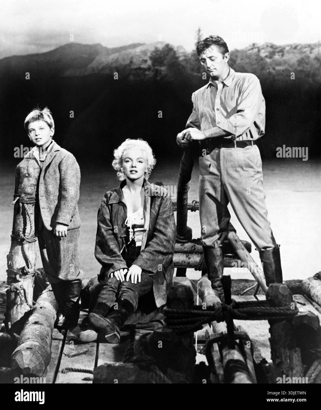 Dieses Vintage-Schwarz-weiß-Foto aus dem Jahr 1954 zeigt Tommy Rettig, Marilyn Monroe und Robert Mitchum in einem Werbefoto für den Film „River of No Return“. Die ikonischen Schauspieler werden auf einem Floß dargestellt, was auf Abenteuer und Drama in der Erzählung des Films hindeutet. Stockfoto