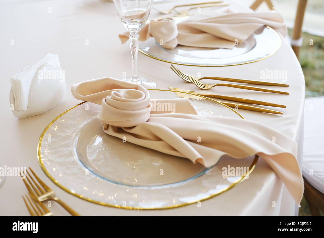 Die Tischdekoration umfasst goldenes Besteck, Gläser und gefaltete Servietten auf weißer Tischdecke mit klaren Tellern und goldenen Rändern, die ordentlich zum Abendessen angeordnet sind Stockfoto