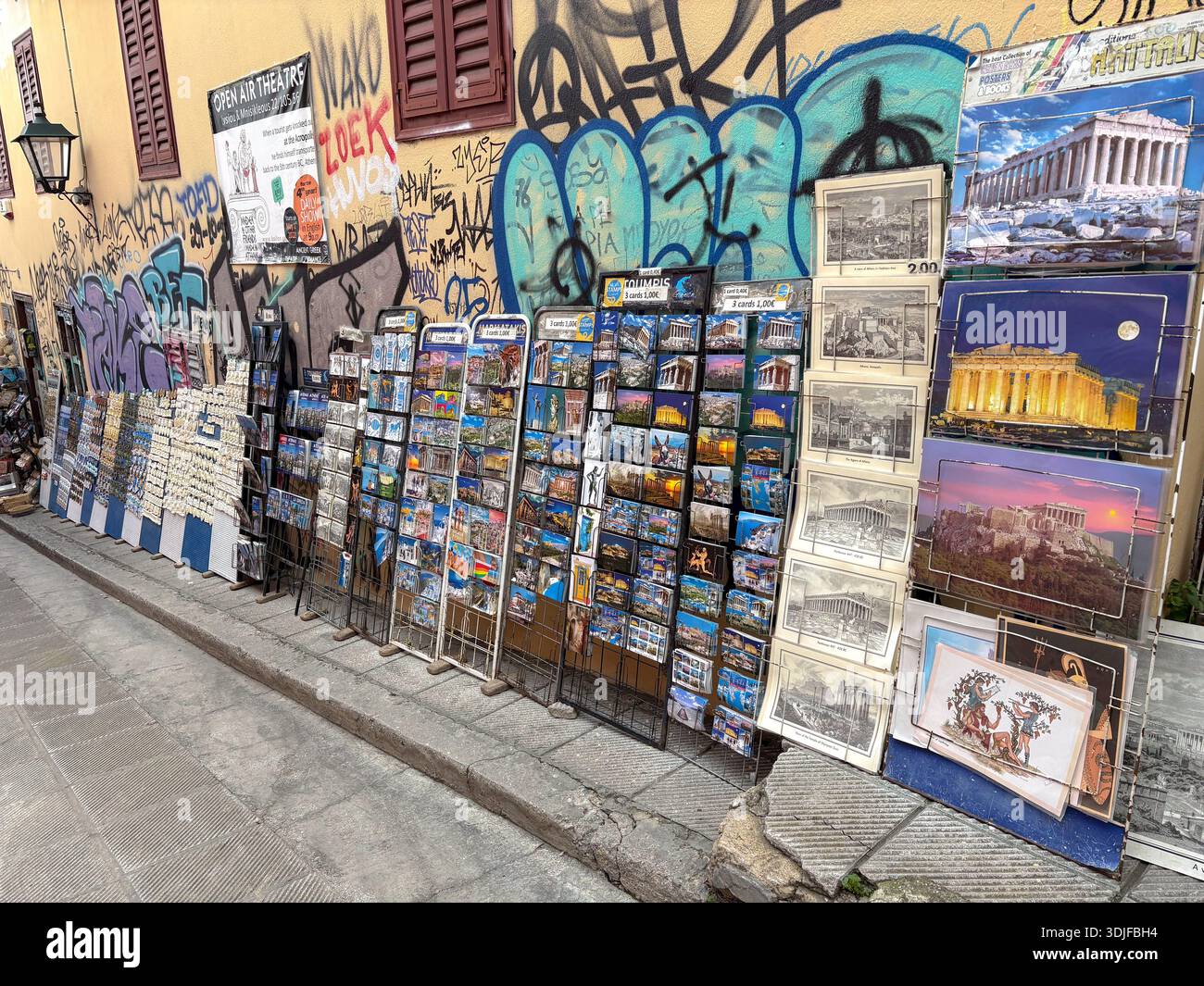 Athen Souvenirstand, der Postkarten mit dem Parthenon verkauft, die an einer hellen Graffiten-Wand in einer historischen Straße gegenübergestellt werden. Stockfoto