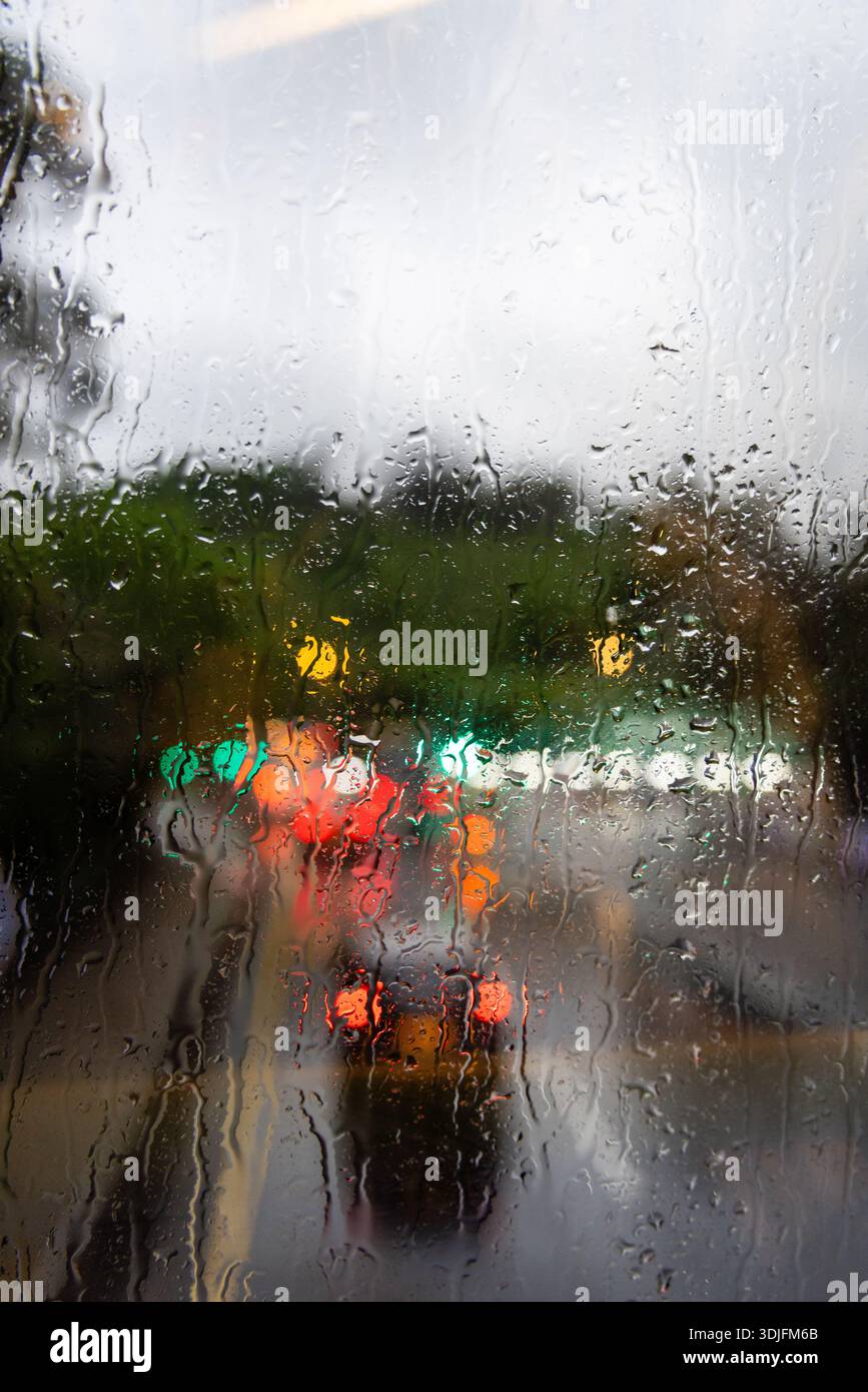 Regentropfen auf Glasfenster. Verschwommene Ampeln im Hintergrund. Stimmungsvolle Atmosphäre. Textur bei nassem Wetter. Filmisches Bokeh. Stockfoto