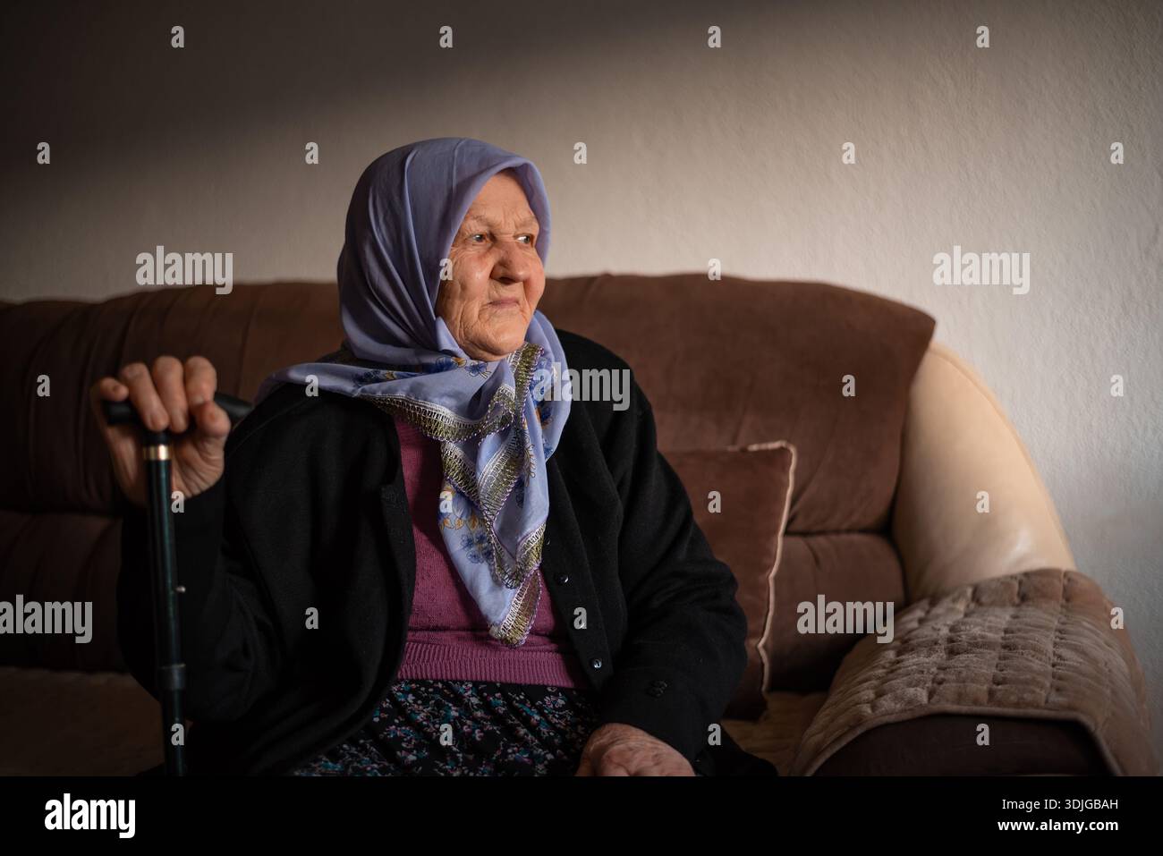 Porträt einer alten muslimischen Frau mit ihrem Gehstock zu Hause, der traditionellen Hijab trägt und wegblickt. Stockfoto