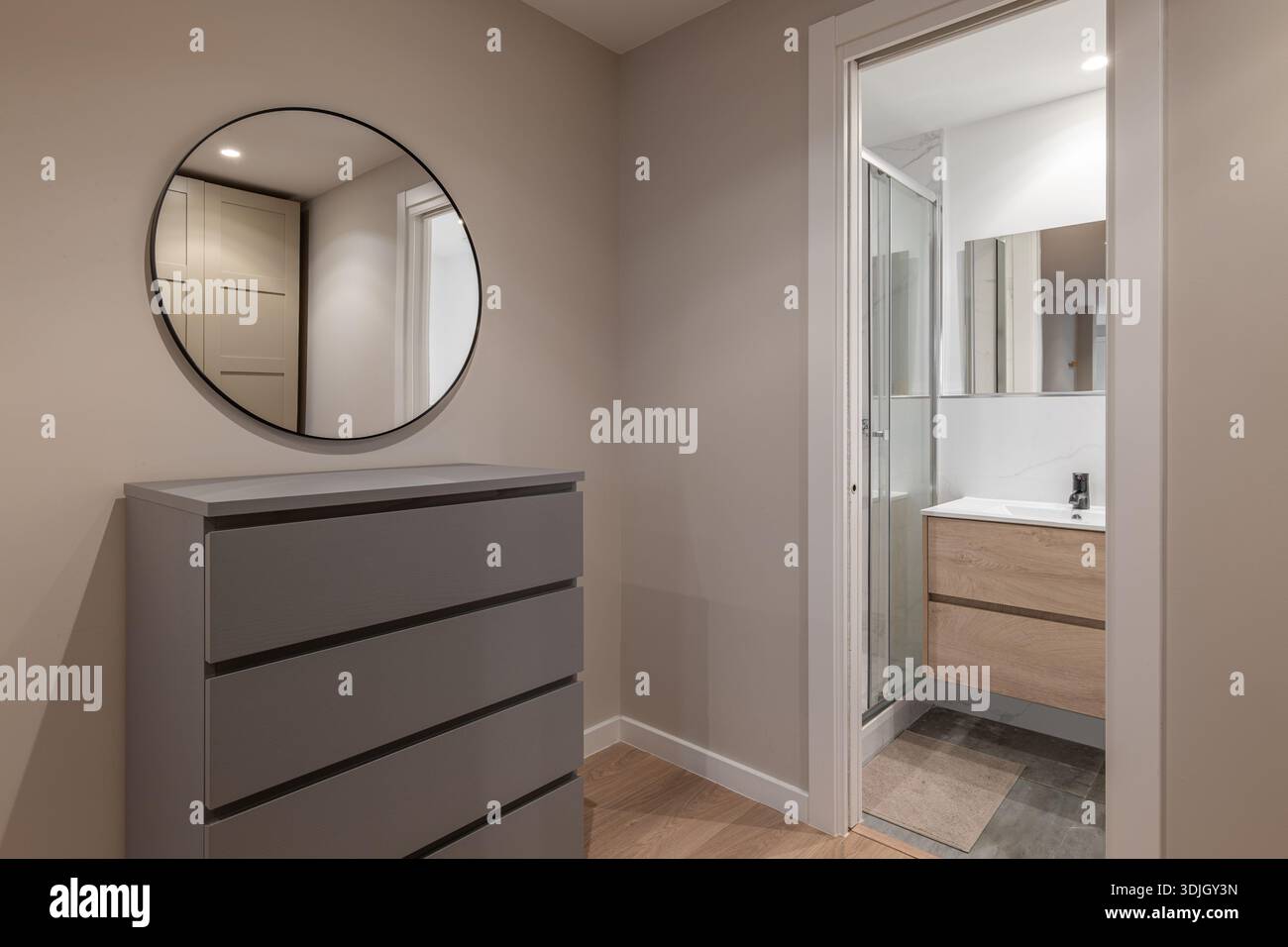 Modernes Badezimmer mit grauer Kommode und rundem Spiegel. Der Raum ist gut beleuchtet und verfügt über eine Glasdusche und einen Holzwaschtisch. Stockfoto