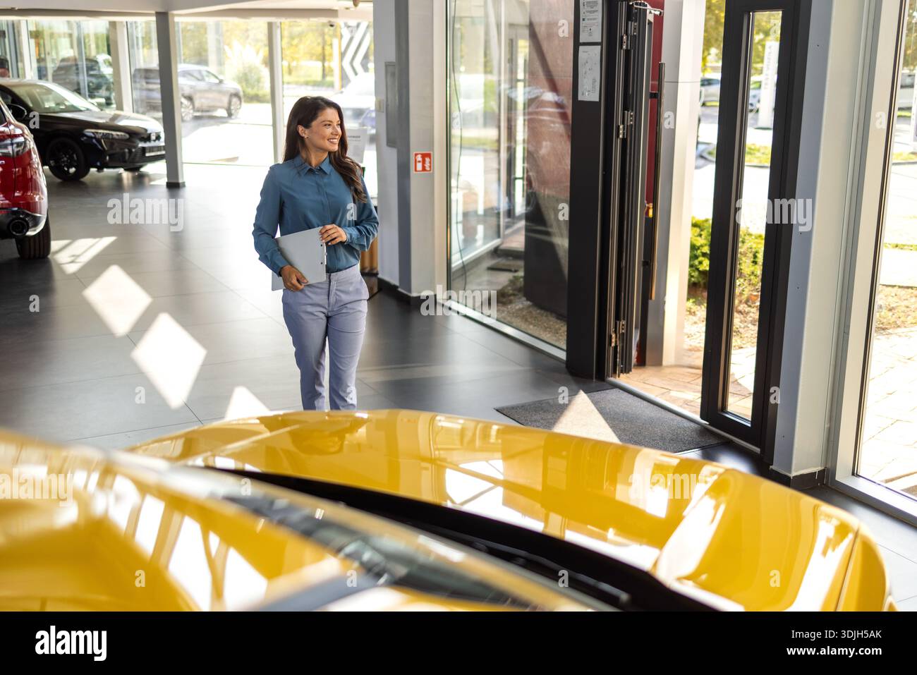 Lächelnde Verkäuferin, die einen Tablet-PC hält, während sie die Kamera im Ausstellungsraum eines neuen Autos betrachtet. Professioneller Autohändler posiert stolz im Autohaus und lächelt zu Stockfoto