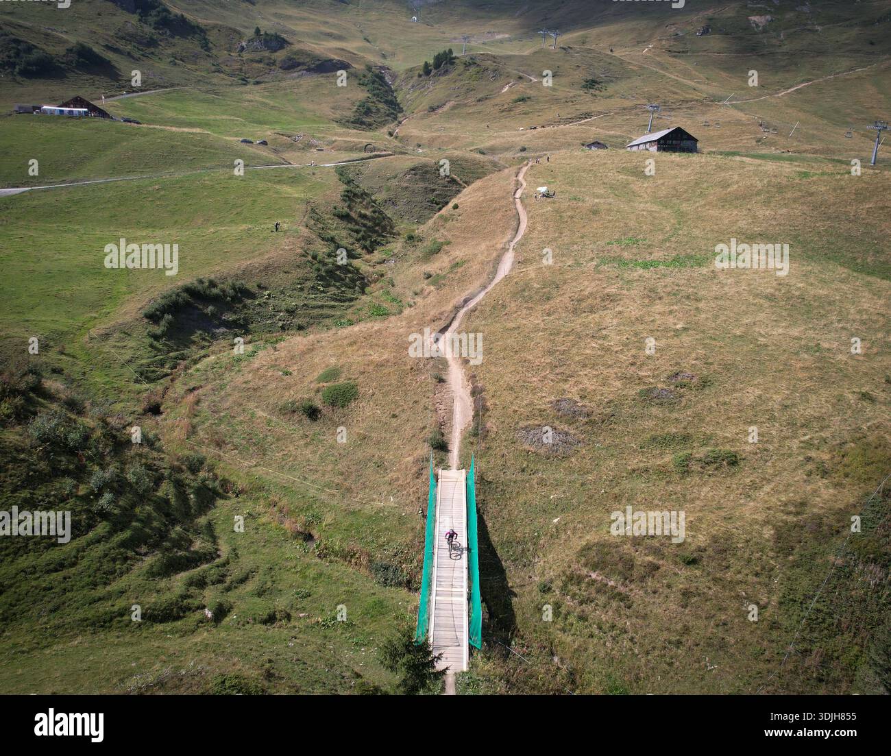 Aus der Vogelperspektive eines einsamen Radfahrers, der eine lebhafte grüne Brücke überquert, inmitten von sanften Hügeln und gewundenen Wegen, Val-d'Illiez, Wallis, Schweiz. Stockfoto