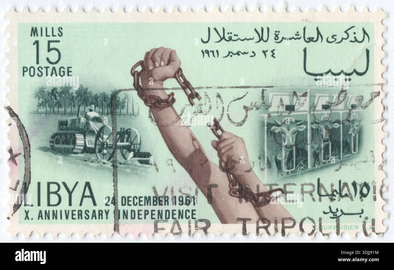 LIBYEN - 24. Dezember 1961: 15 Milliemer blassgrüne, grüne und braune Briefmarke erinnert an den 10. Jahrestag der libyschen Unabhängigkeit und feiert das Jahrzehnt seit der Vereinigung von Tripolitanien, Kyrenaika und Fezzan 1951 zu einem souveränen Königreich. Das zentrale Design zeigt ein symbolisches Bild von Händen, die eine schwere Metallkette brechen und das Ende der italienischen Kolonialherrschaft und die Erlangung nationaler Freiheit darstellen. Im Hintergrund geht die Szene zu Themen des Friedens und Fortschritts über, wobei Rinder auf einer fruchtbaren Weide weiden und ein moderner Traktor das Land bebaut Stockfoto