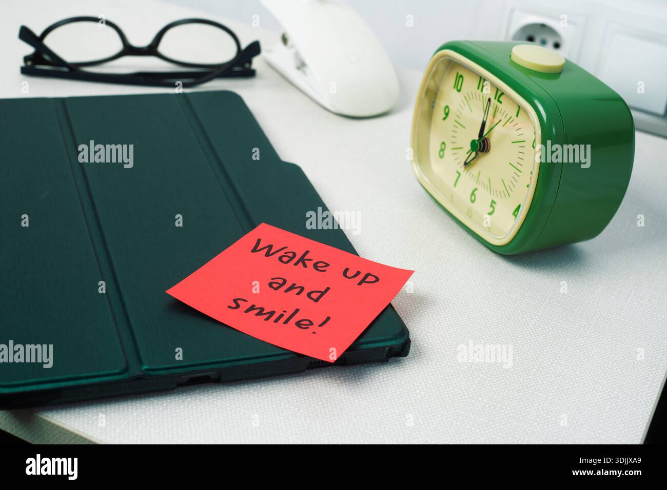 Auf einem weißen Nachttisch befindet sich neben einem Tablet und einer Brille ein grüner Wecker, während eine rote Haftnotiz aufwacht und ein Lächeln auf dem Gerät aufliegt Stockfoto