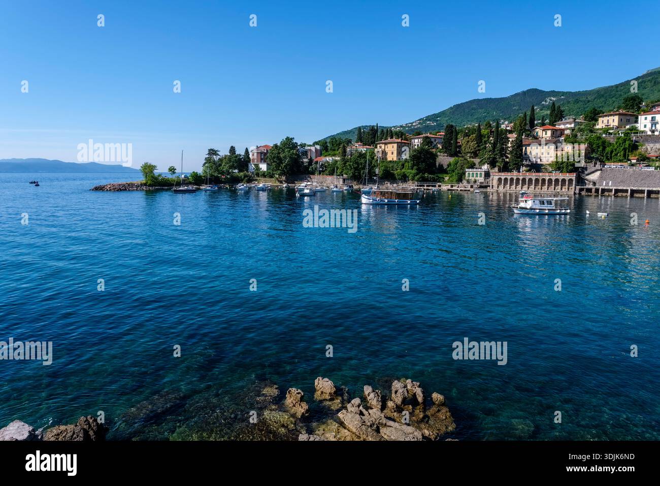 Boote liegen im Hafen von IKA. Küstengebäude und üppige grüne Hügel unter einem klaren blauen Himmel in der Ferne. Stockfoto