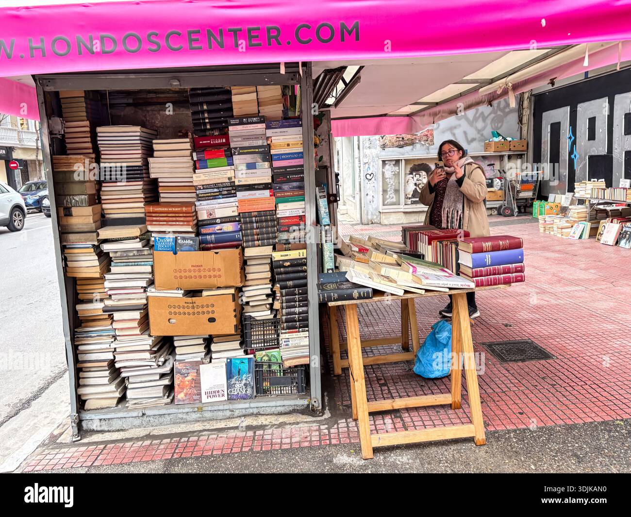Bücherstand und Verkäufer auf einer Straße in Athen gebraucht. Straßenverkäufer, der gebrauchte Bücher von einer Kioskstruktur verkauft, die häufig auf griechischen Straßenmärkten zu finden ist. Stockfoto