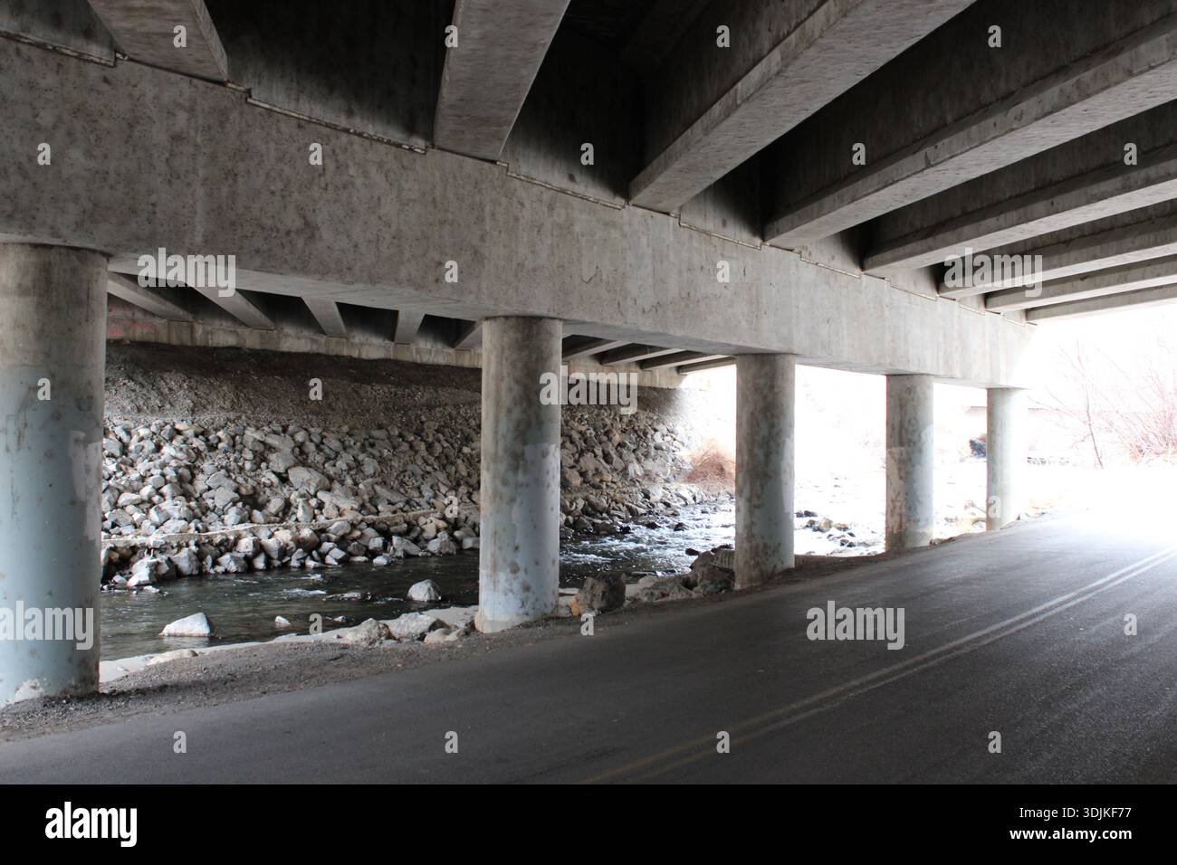 Leerer Fußgängerweg unter einer städtischen Straße mit starker linearer Perspektive, Betonsäulen und einem schmalen Bach in ruhiger Winterumgebung. Stockfoto