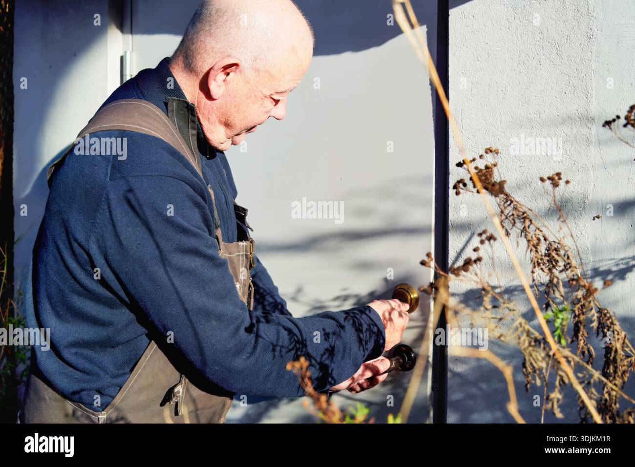 Ein älterer Mann in Arbeitskleidung öffnet eine Tür mit einem Messingknopf, umgeben von der Natur, und veranschaulicht den Moment des Betretens einer neuen Umgebung Stockfoto