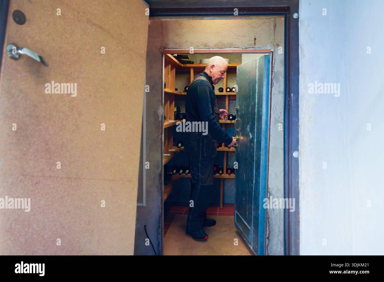 Ein Mann in schwarzen Overalls öffnet eine blaue Tür zu einem Weinlagerbereich, enthüllt Regale mit Flaschen, die eine Atmosphäre der Vorfreude schaffen Stockfoto