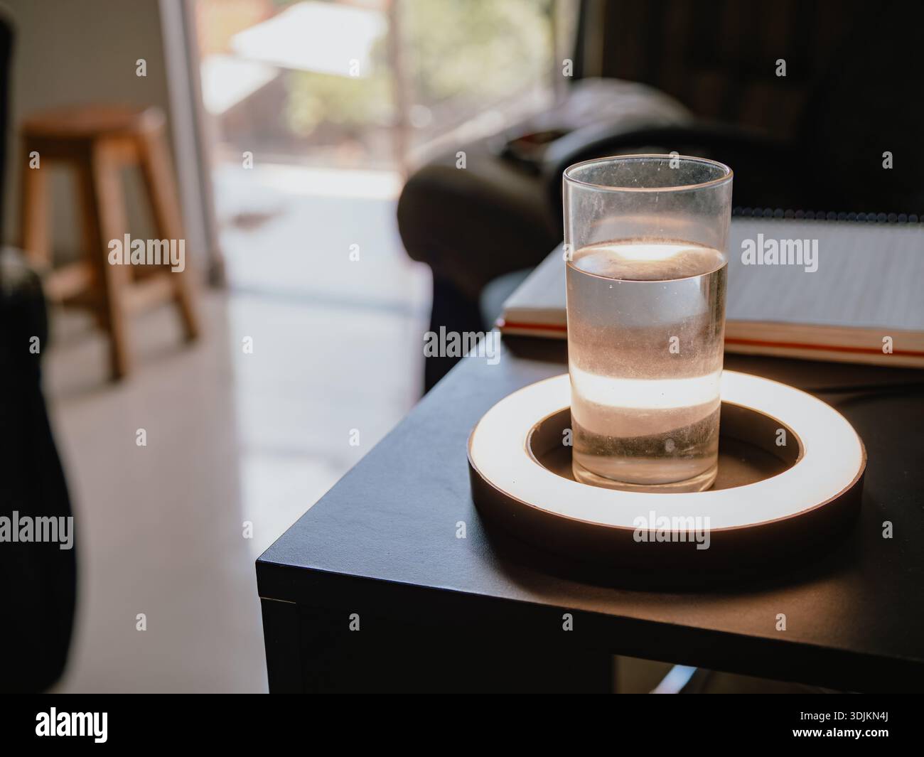 Foto eines Glasglases mit Wasser auf einem schwarzen Tisch, beleuchtet durch warmes Licht, mit einem unscharfen Innenhintergrund. Ideal zur Hydration, hea Stockfoto