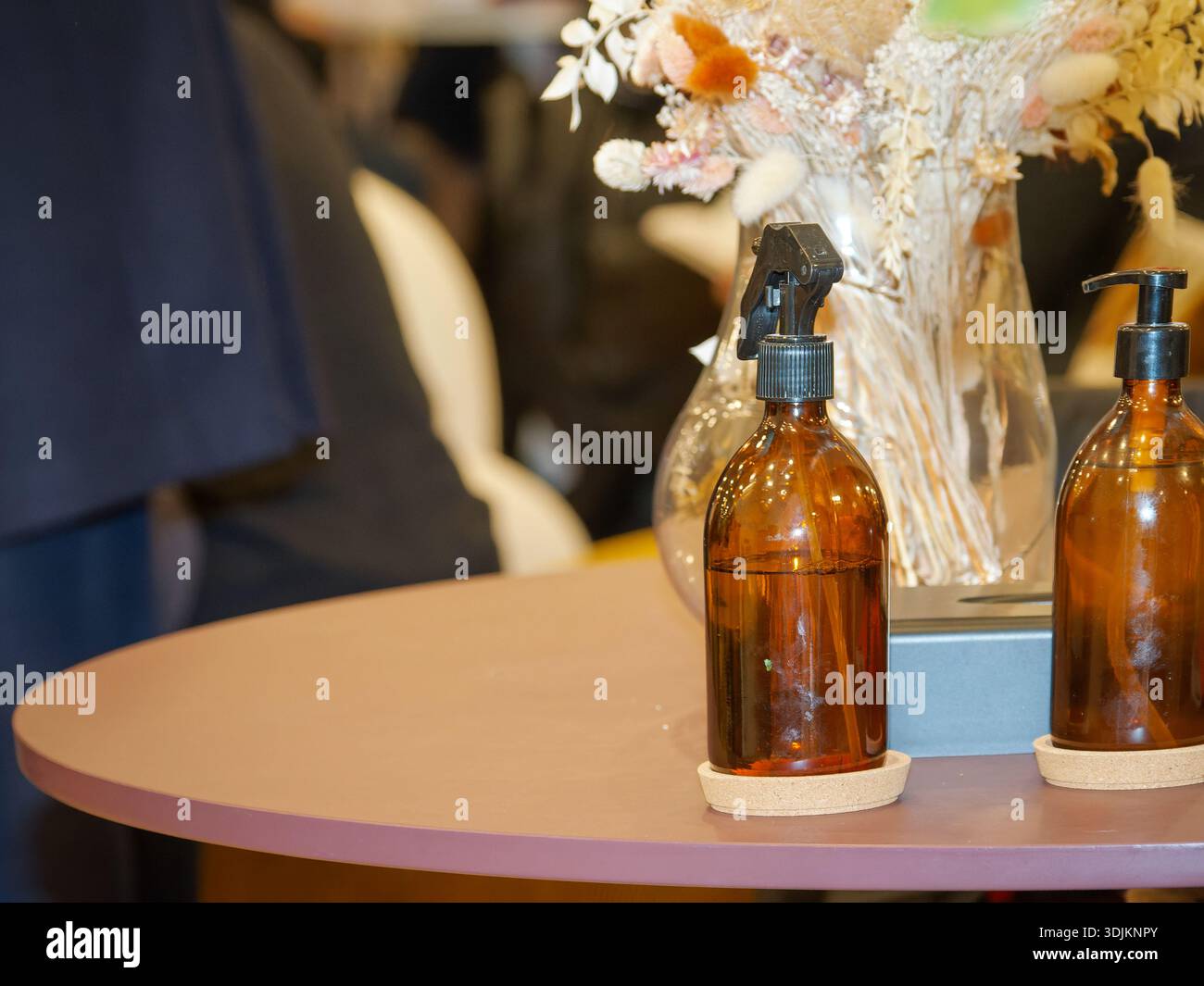 Sprühflaschen aus braunem Glas auf einem kleinen runden Tisch mit Blumendekor und minimalistischen Details im Innenraum, die auf Reinigung oder Kosmetikprodukte hinweisen Stockfoto
