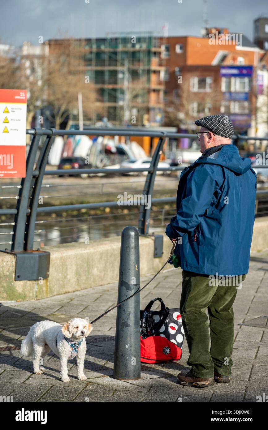 Ein älterer Herr mit einem kleinen Terrierhund, der Boote und Schiffe in Portsmouth Harbour, Hampshire, Großbritannien, ansieht Stockfoto