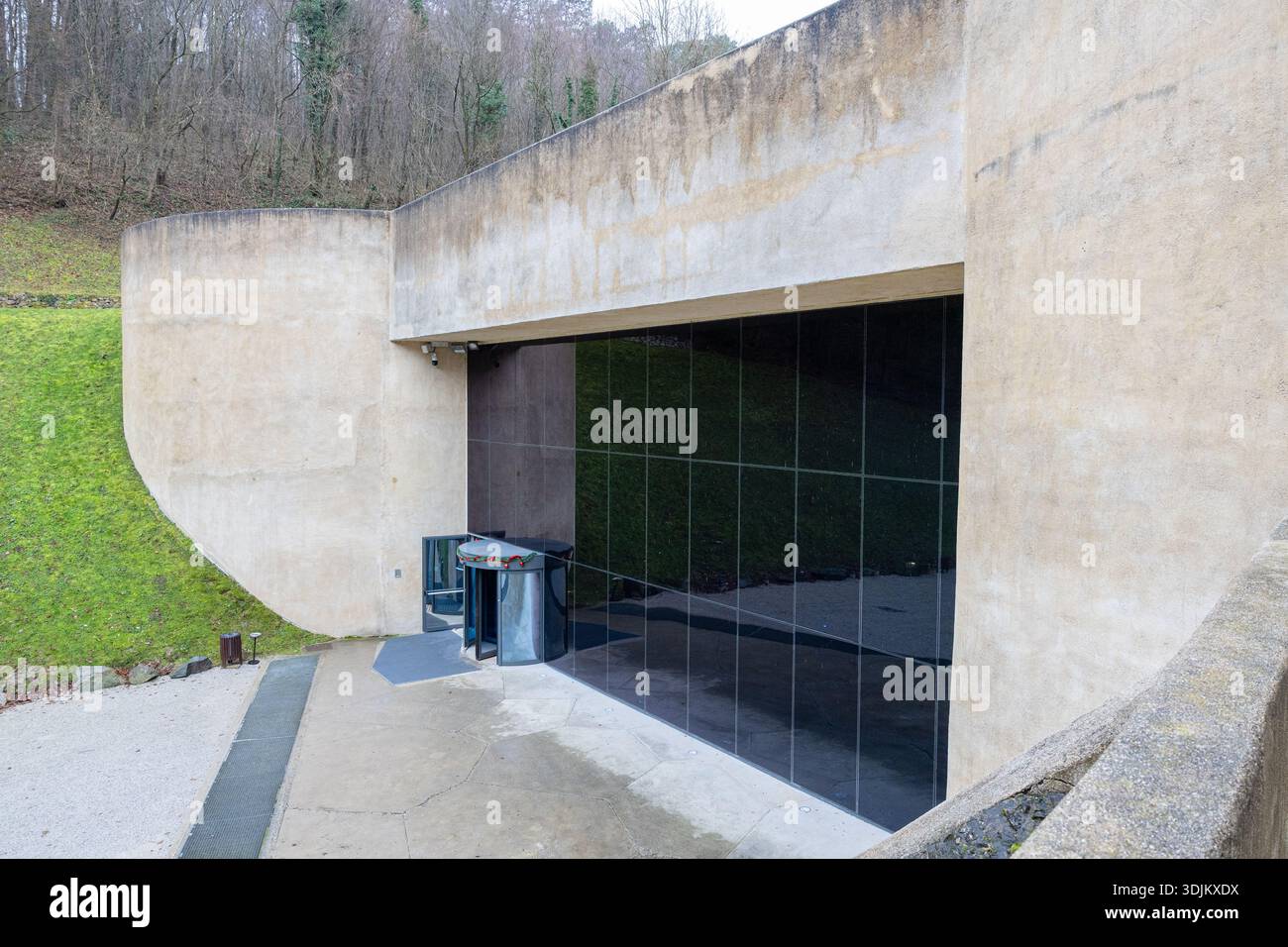 KRAPINA, KROATIEN, 01.02.2026: Eine markante Betonwand umrahmt eine große schwarze Glastür in einem Park im Freien. Reflektierende Einsätze, minimalistisches Design Stockfoto