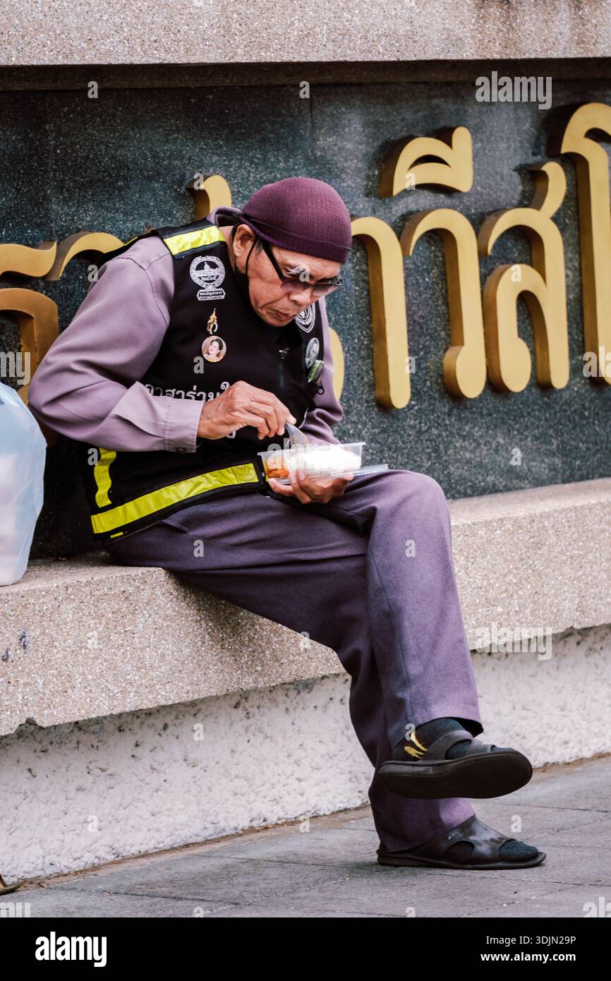 Bangkok, Thailand, 19. November 2025, sitzt ein einheimischer älterer Mann lässig auf einem Steinvorsprung und genießt seinen Snack vor einer lebhaften Kulisse thailändischer Buchstaben Stockfoto