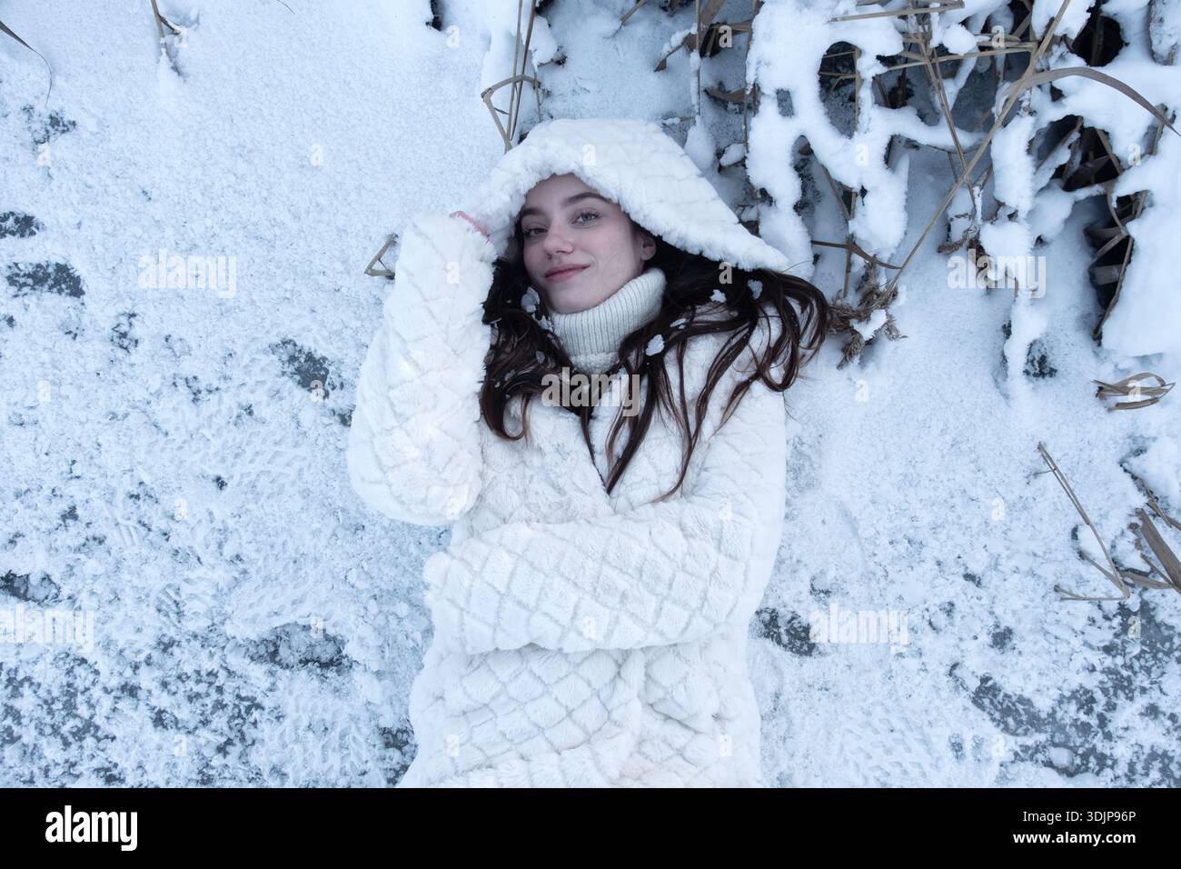 Junge Frau, die im Neuschnee liegt und einen weißen Wintermantel und eine Kapuze trägt, die entspannt und zufrieden von oben aussieht und Wärme, Intimität, Kontrast und Qu ausdrückt Stockfoto