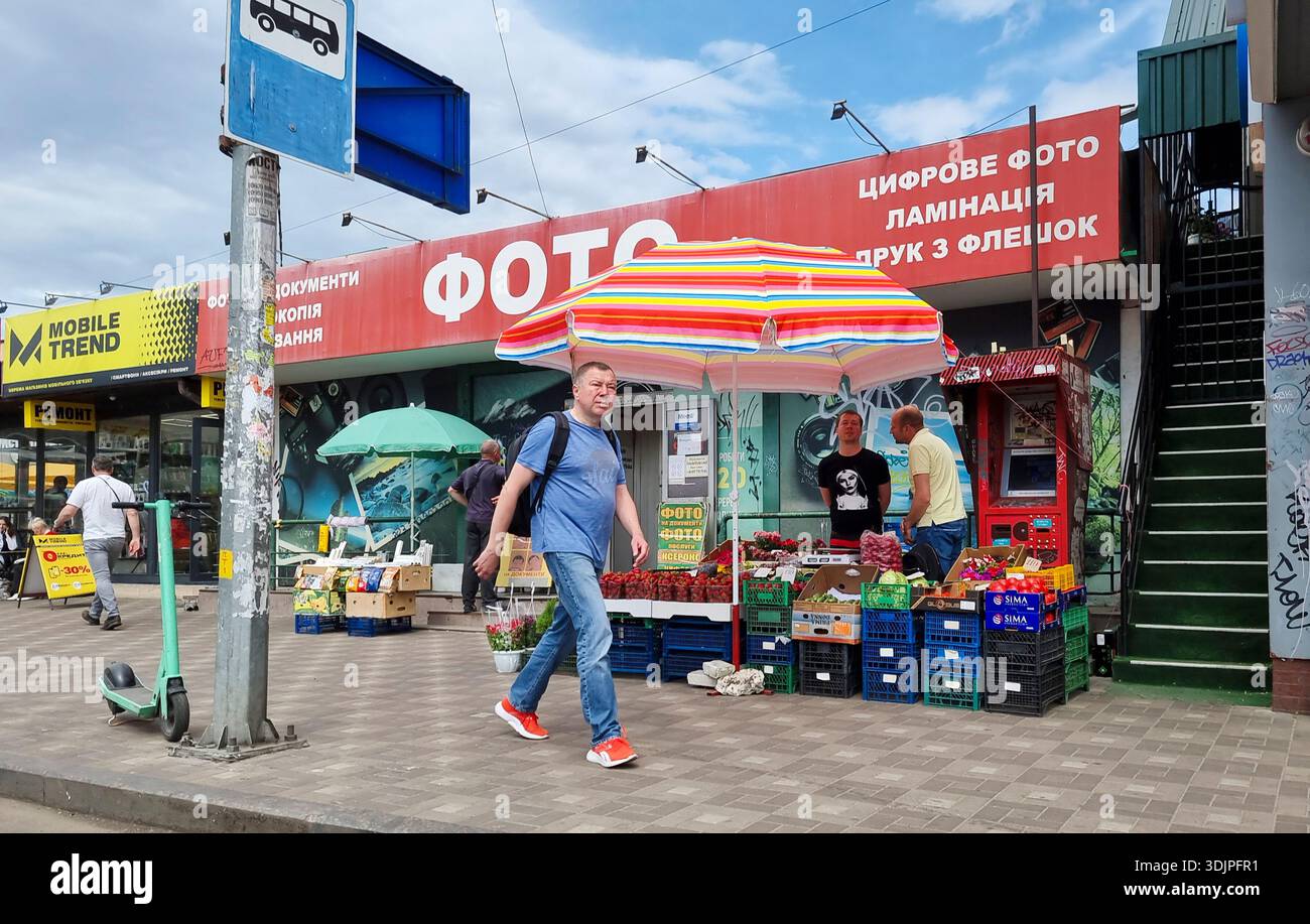 Kiew, Ukraine - 6. Mai 2023 : ein Mann, der an einem farbenfrohen Kiewer Obst- und Gemüsestand an einer Bushaltestelle und Schaufensterfronten vorbeiläuft und den alltäglichen städtischen mar erfasst Stockfoto