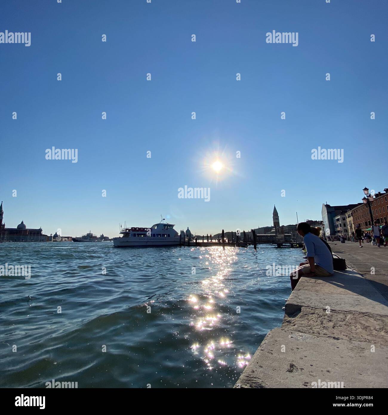 Frau, die nachdenklich auf einem Kanalpier von Venedig sitzt und vorbeifahrende Boote und den Canale Grande mit dem Markusdom unter einem sonnigen blauen Himmel ansieht. - Smartphone-aufgenommenes Stockfoto