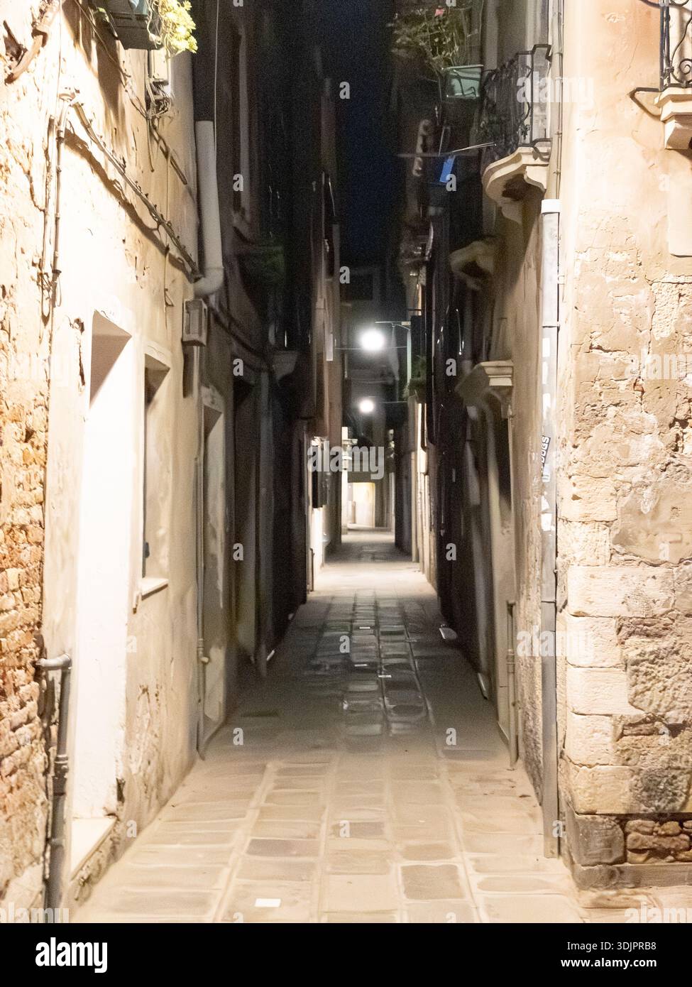 Stimmungsvolle enge Kopfsteinpflasterallee in Venedig bei Nacht, beleuchtet von sanften Straßenlaternen, die lange Schatten auf verwitterten Steinmauern und b - Smartphone-aufgenommenes Stockfoto