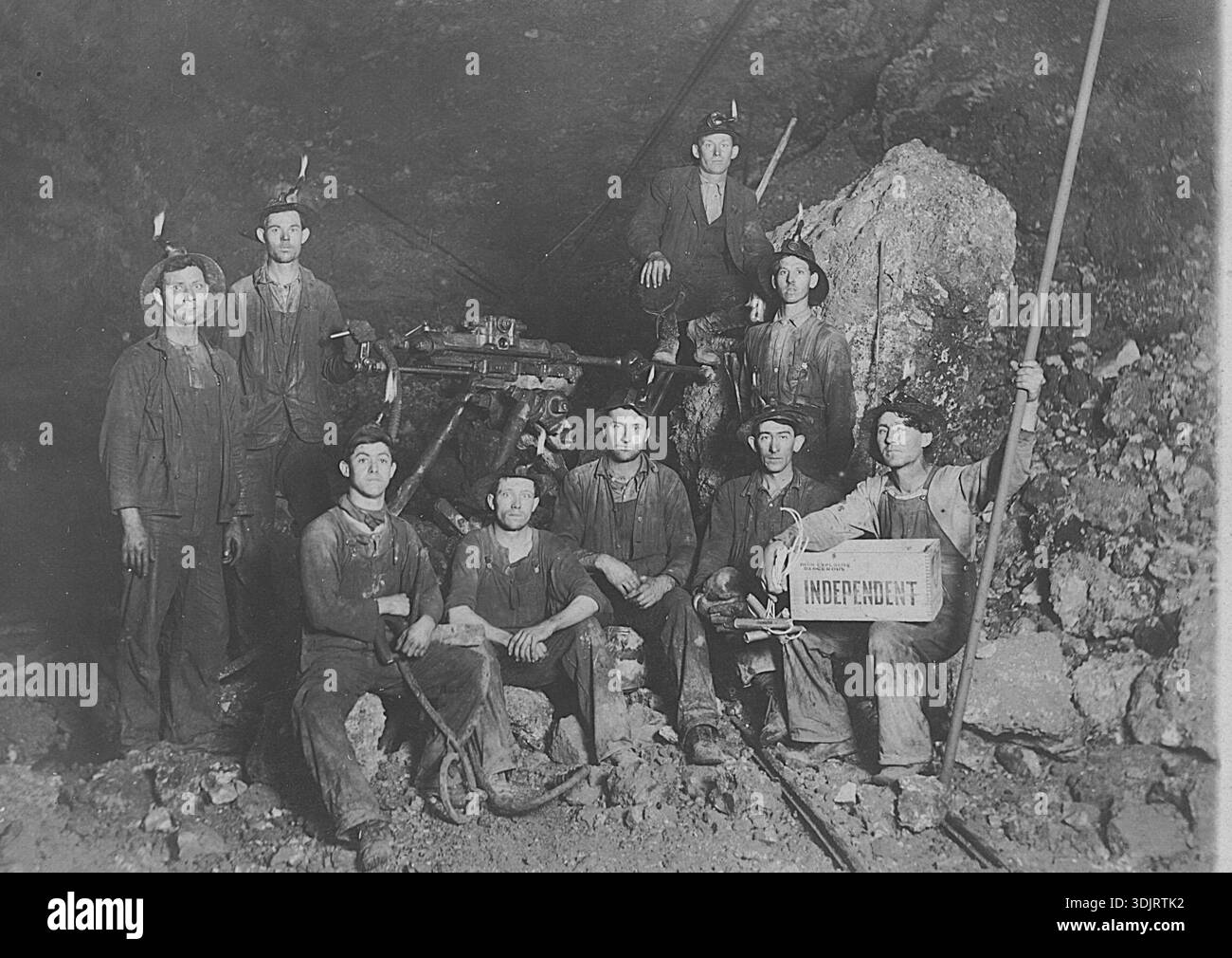 Neun Kohlebergleute posieren mit ihrem hydraulischen Bohrer, der tiefe Löcher bohrt, in die die Bergleute Dynamit schieben. Ein Mann hat eine offene Kiste Dynamit auf seinem Schoß. Sie trägt die Aufschrift „High explosive Dangerous Independent“. Alle Männer tragen Helme mit Öllampen. Die Helme haben längere Rückenkrempe, um Steine zu halten und Wasser vom Kopf zu tropfen. Beachten Sie die Laufschienen unten rechts. Um meine anderen Bergbaufotos zu sehen, suchen Sie nach Prestor Vintage Minenbohrer Stockfoto