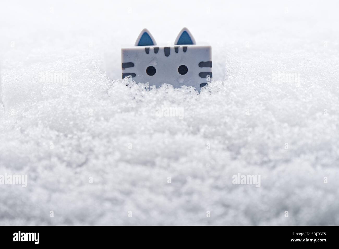 Niedliche katzenförmige Figur, die aus frischem Schnee blickt, minimalistische Winterszene mit sanftem Licht und verspielter Stimmung. Stockfoto