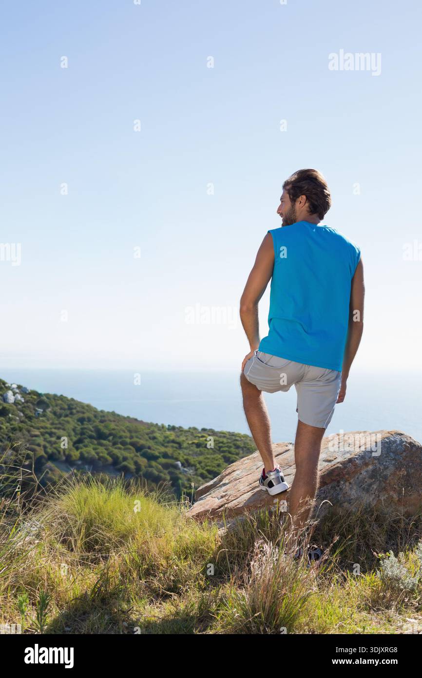 Der Mensch steht mit einem Fuß auf dem Felsen und blickt auf das ruhige blaue Meer vom Küstenhang. Wanderer, Landschaft, Küste, Aussichtspunkt, Natur, Einsamkeit, Sonnenschein Stockfoto