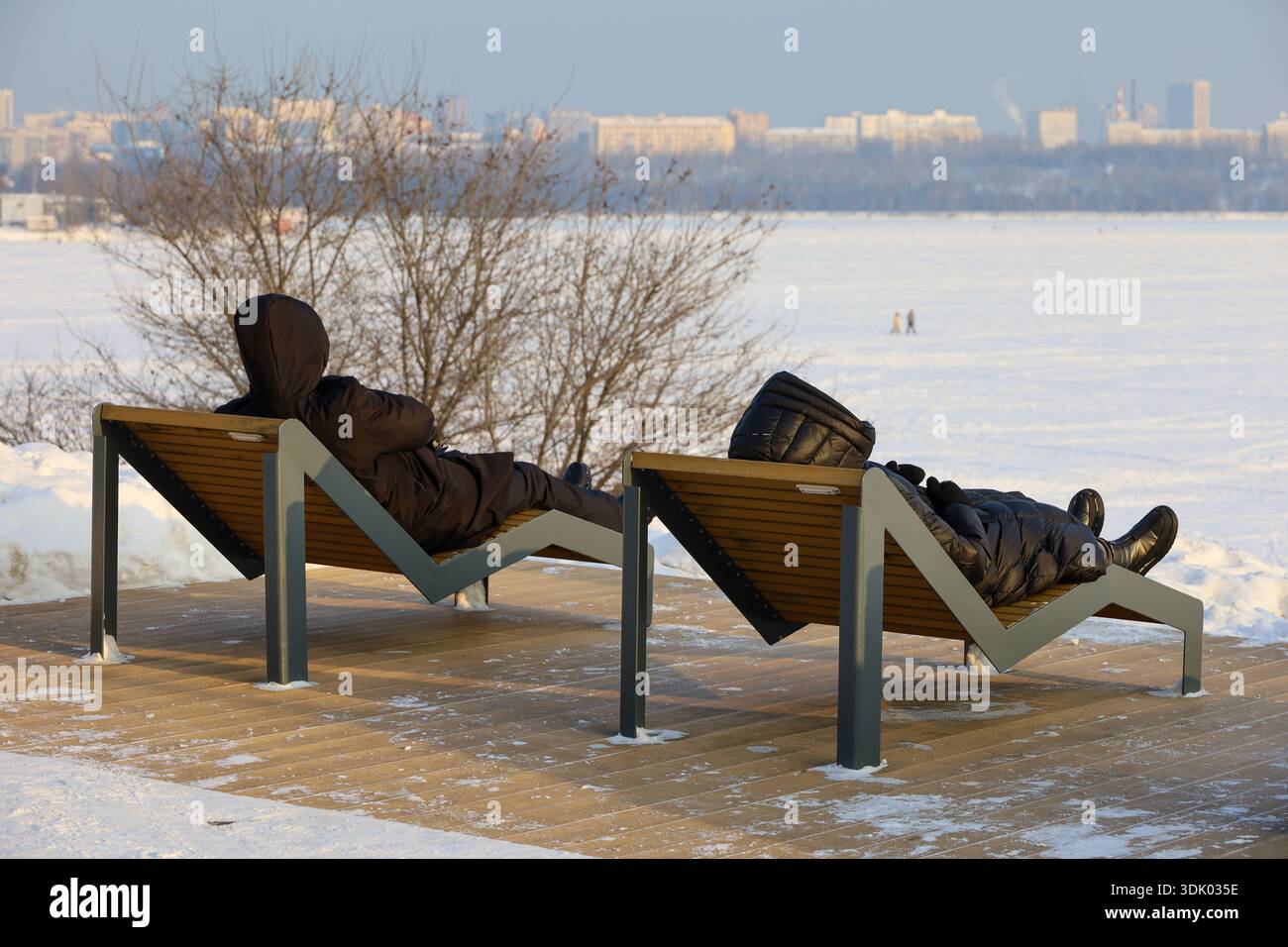 Leute in warmen Kleidern liegen in Liegestühlen am Winterstrand vor dem Hintergrund der Stadt. Urlaub bei kaltem Wetter Stockfoto