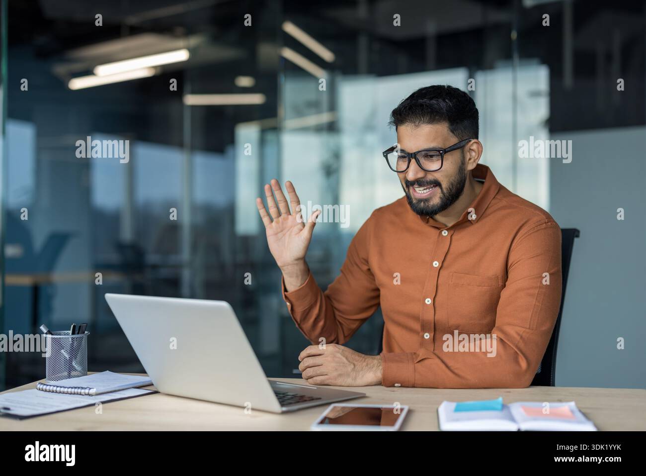 Junger Geschäftsmann, der von seinem modernen Büro aus an einer Videokonferenz teilnimmt, lächelt und auf einem Laptop Kollegen oder Kunden zuwinkt und dabei die Aufmerksamkeit von Stockfoto