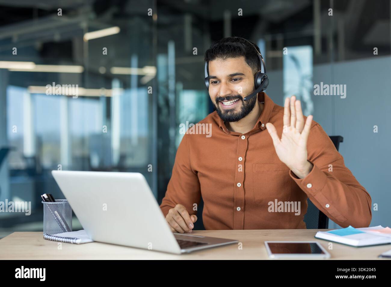 Selbstbewusster bärtiger Mann, der ein Headset trägt und lächelt, an einem Schreibtisch in einem modernen Büro sitzt und aktiv an einem erfolgreichen virtuellen Bus teilnimmt Stockfoto