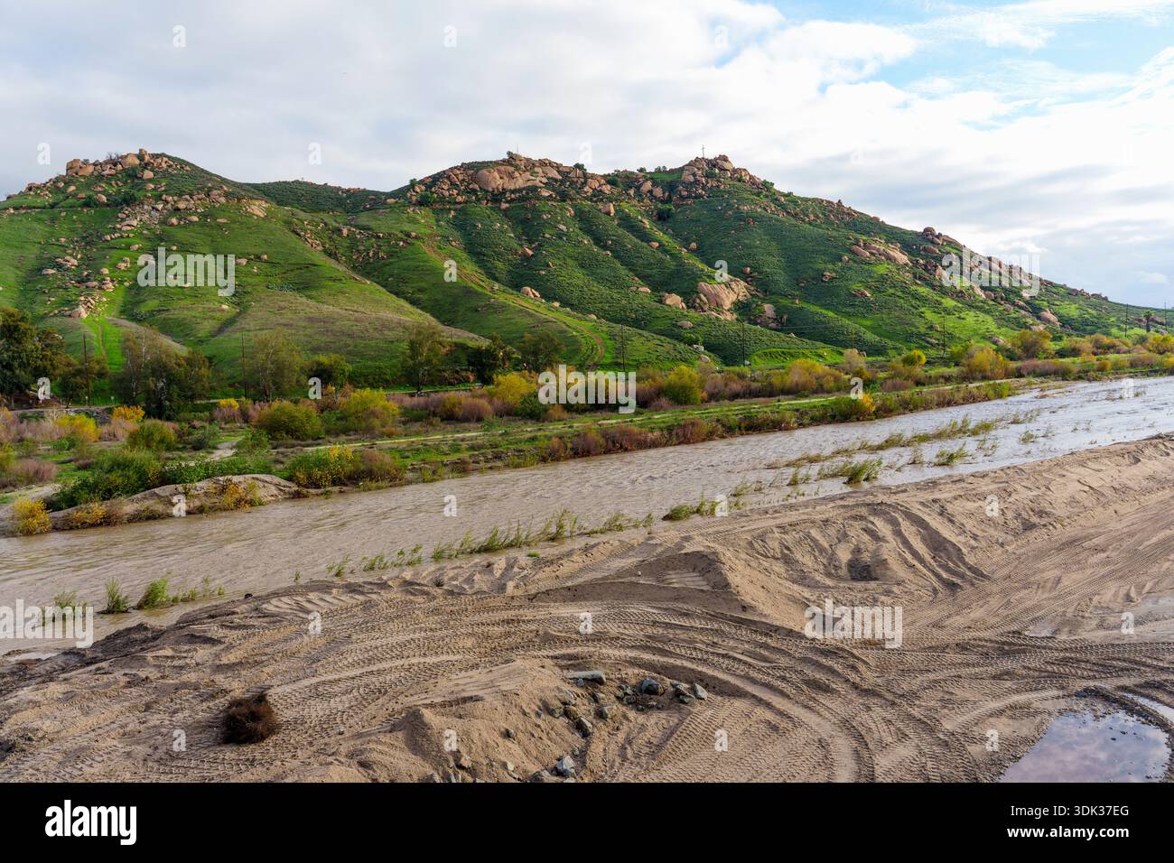 Der schlammige Fluss fließt an üppigen grünen Hügeln vorbei, die mit Felsen und verstreuten Bäumen bedeckt sind, unter einem bewölkten Himmel in der Gegend um Frank A. Miller Mount Rubidoux. Stockfoto