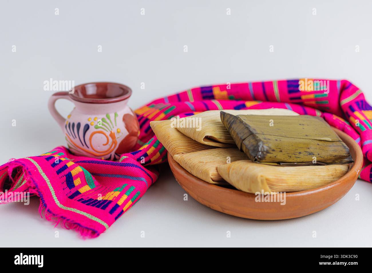 Traditionelles mexikanisches Frühstück mit verschiedenen Tamales und einer dekorierten Tontasse auf rosafarbener Tischdecke und weißem Hintergrund Stockfoto