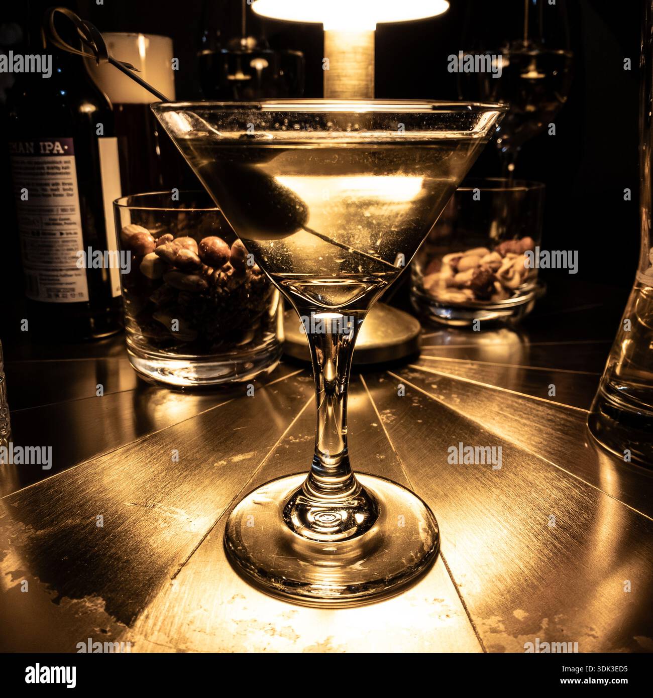 Klassischer schmutziger Wodka Martini in einem Martini-Glas, garniert mit Olivenspieß, umgeben von Nussschalen unter warmem Lampenlicht auf einer metallischen Goldoberfläche - Smartphone-aufgenommenes Stockfoto
