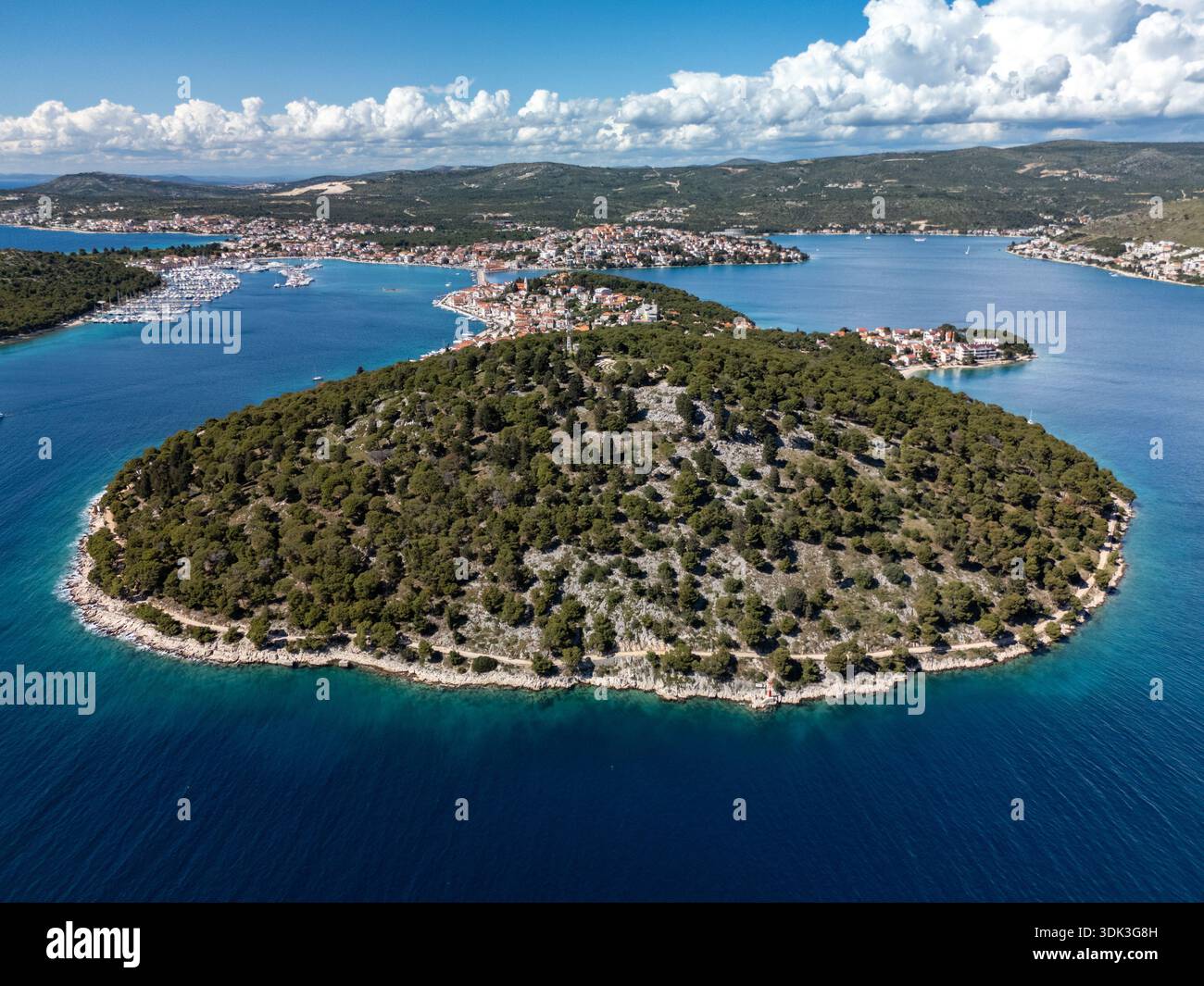 Aus der Vogelperspektive auf die üppige, grüne Insel, umgeben vom tiefblauen Meer, im Kontrast zum klaren Himmel und der fernen Küstenstadt Rogoznica, Kroatien. Stockfoto