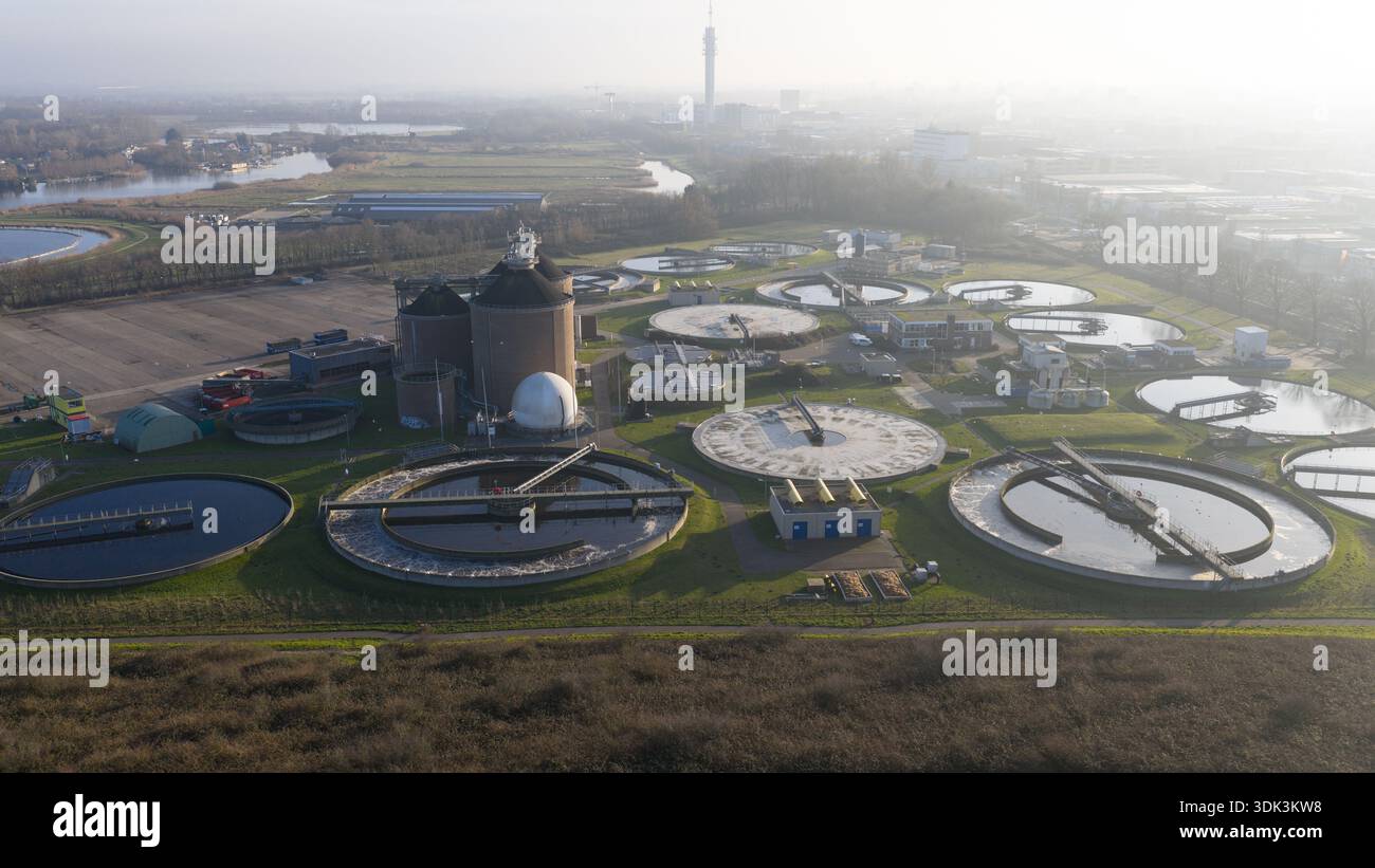 Aus der Vogelperspektive der kreisförmigen Wasseraufbereitungsbecken, die den Himmel reflektieren, neben zylindrischen Strukturen, die vor einem trüben Horizont stehen, Abwasserbehandlung Stockfoto