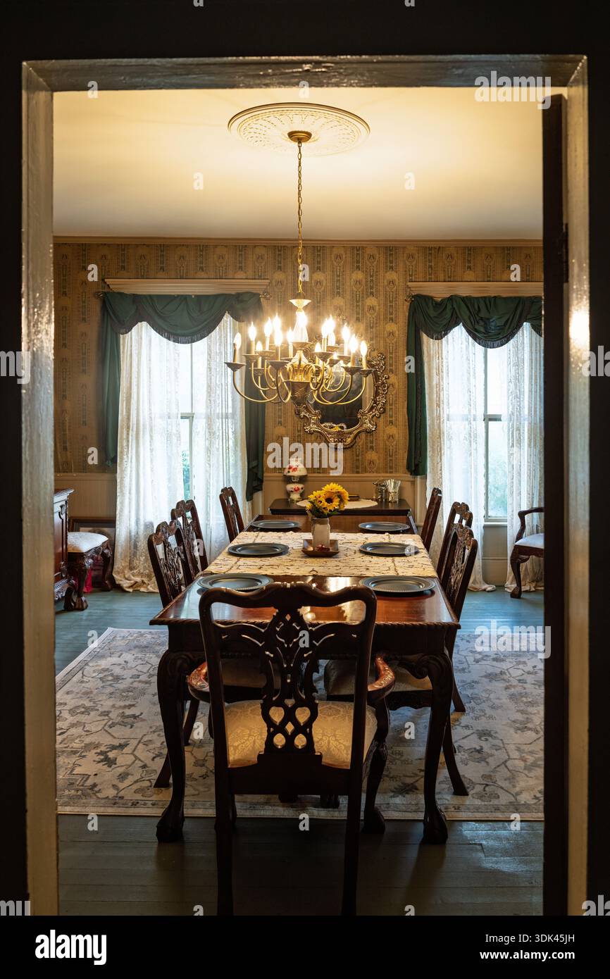 Aus den Schatten eines verdunkelten Flurs wartet ein formeller Speisesaal in der Ruhezeit. Das warme Leuchten eines großen Kronleuchters beleuchtet einen Tisch, für den er gedeckt ist Stockfoto
